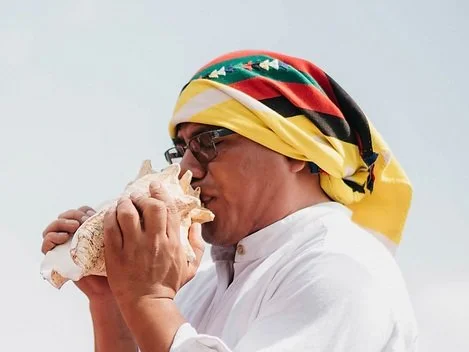 Man with glasses wearing a colorful headscarf, holding and kissing a large piece of bread.