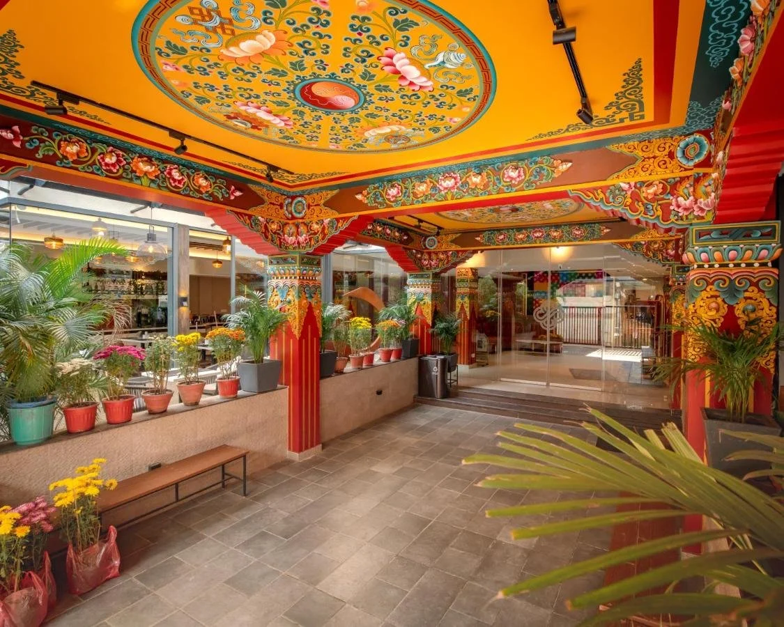 Colorful, ornate ceiling with traditional Asian patterns and floral designs in a restaurant or cultural space. Potted plants line the edges, and the space features glass doors and windows, with a tiled floor.