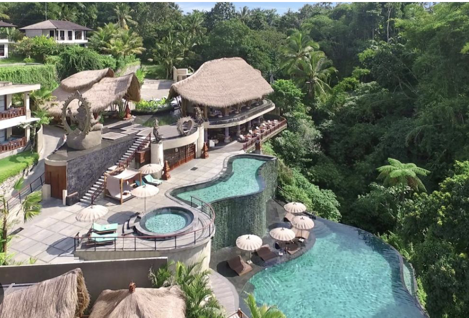 A luxurious infinity pool at a tropical resort surrounded by lush greenery and thatched-roof structures with poolside loungers and umbrellas.