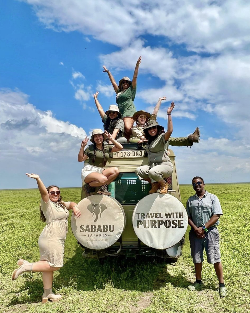 Group of people posing on and around a safari vehicle in an open grassy field, under a sky with clouds. They are smiling, wearing hats, and casual outdoor clothing, with some extending their arms upward and making peace signs.