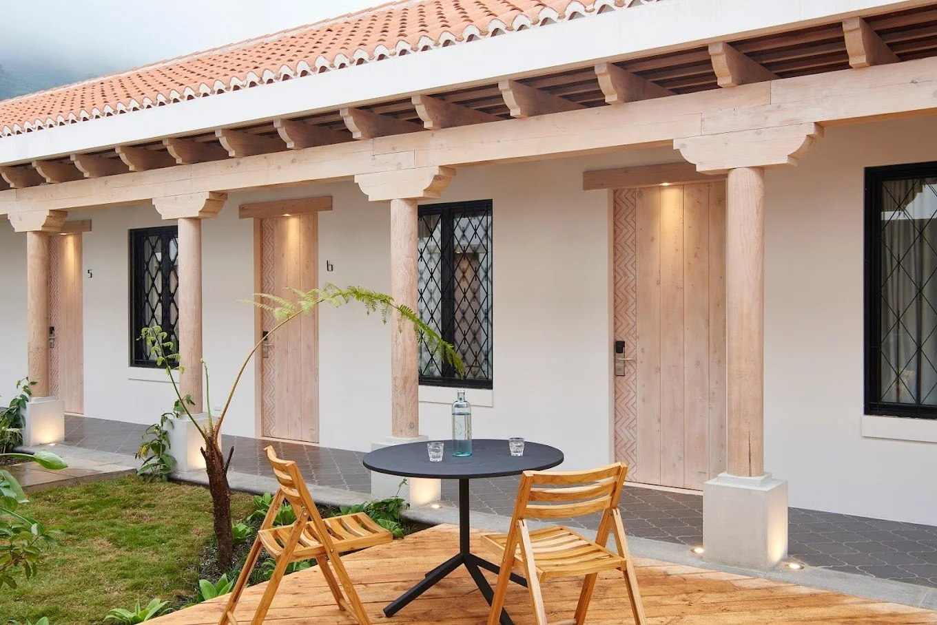 Exterior view of a house with a patio, wooden pillars, and a terracotta tiled roof. There is a small outdoor table with two wooden chairs on a wooden deck, with a bottle and two glasses on the table. Plants and grass are in the foreground.