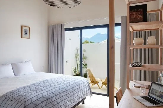 A modern bedroom with a large bed, glass sliding door leading to a balcony with outdoors view, a small table with a laptop, and floating shelves with decor.