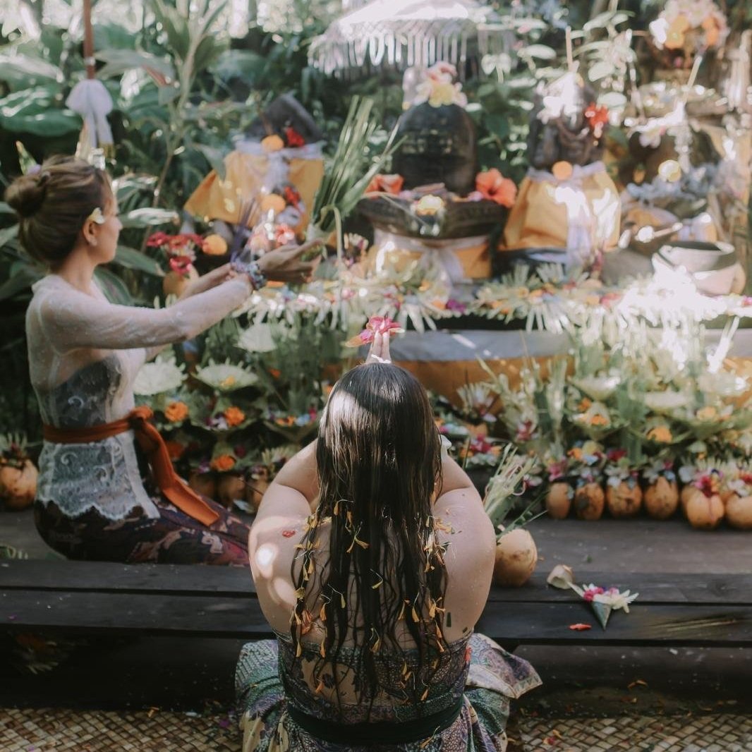 A woman with long wet hair sitting on the ground with her back to the camera, participating in a Balinese cremation ceremony, with an intricately decorated funeral pyre and offerings adorned with flowers and bamboo in the background.