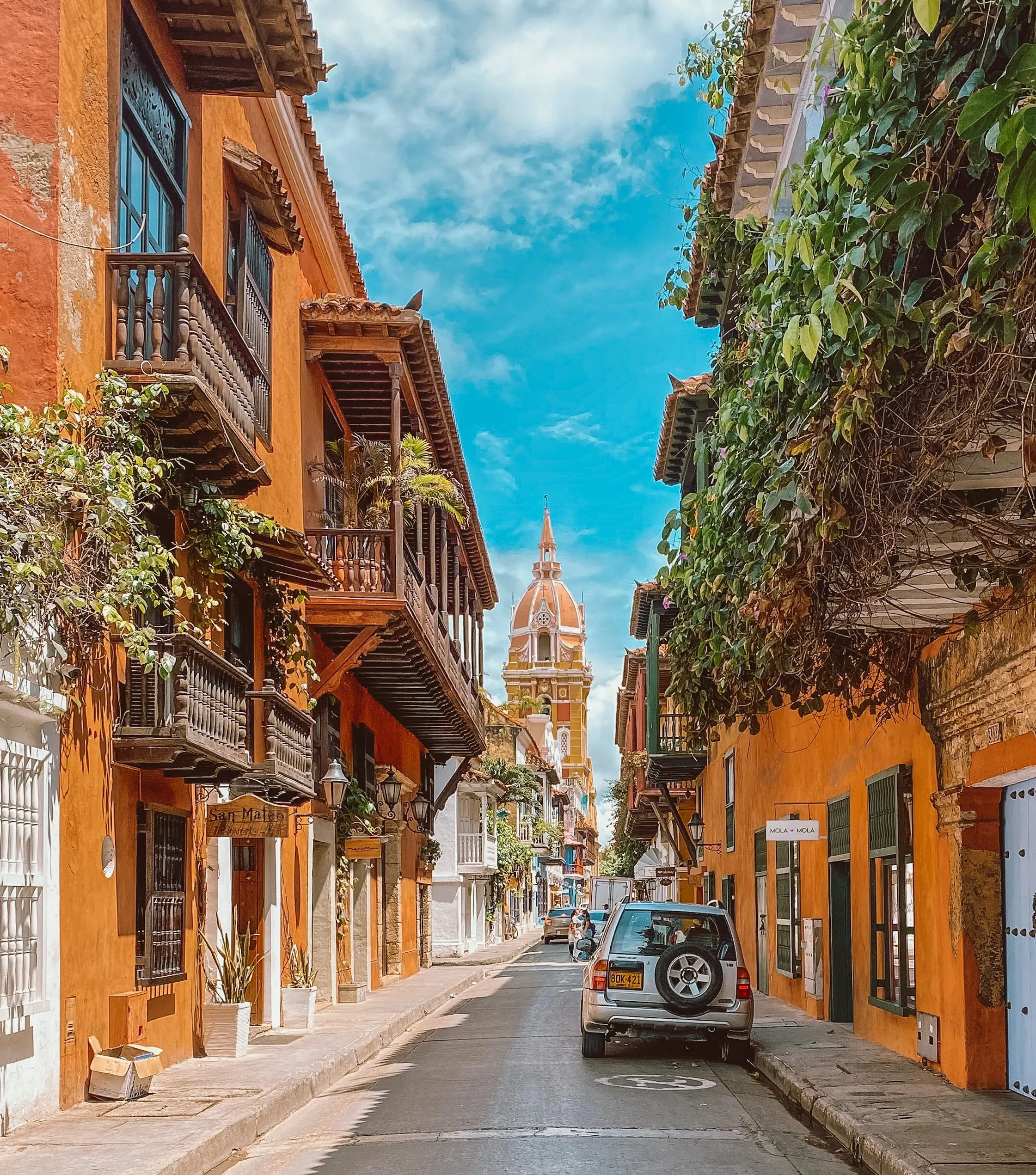 Colorful street view with orange buildings, wooden balconies, lush greenery, and a church tower against a blue sky.