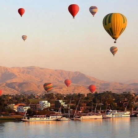 Multiple hot air balloons floating above a river with boats and mountains in the background.