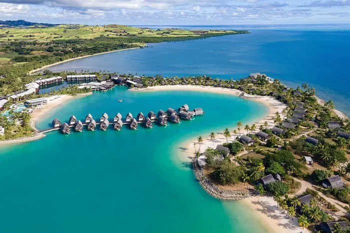Aerial view of overwater bungalows on a tropical lagoon with a sandy beach, surrounded by lush greenery and ocean, with distant land in the background.