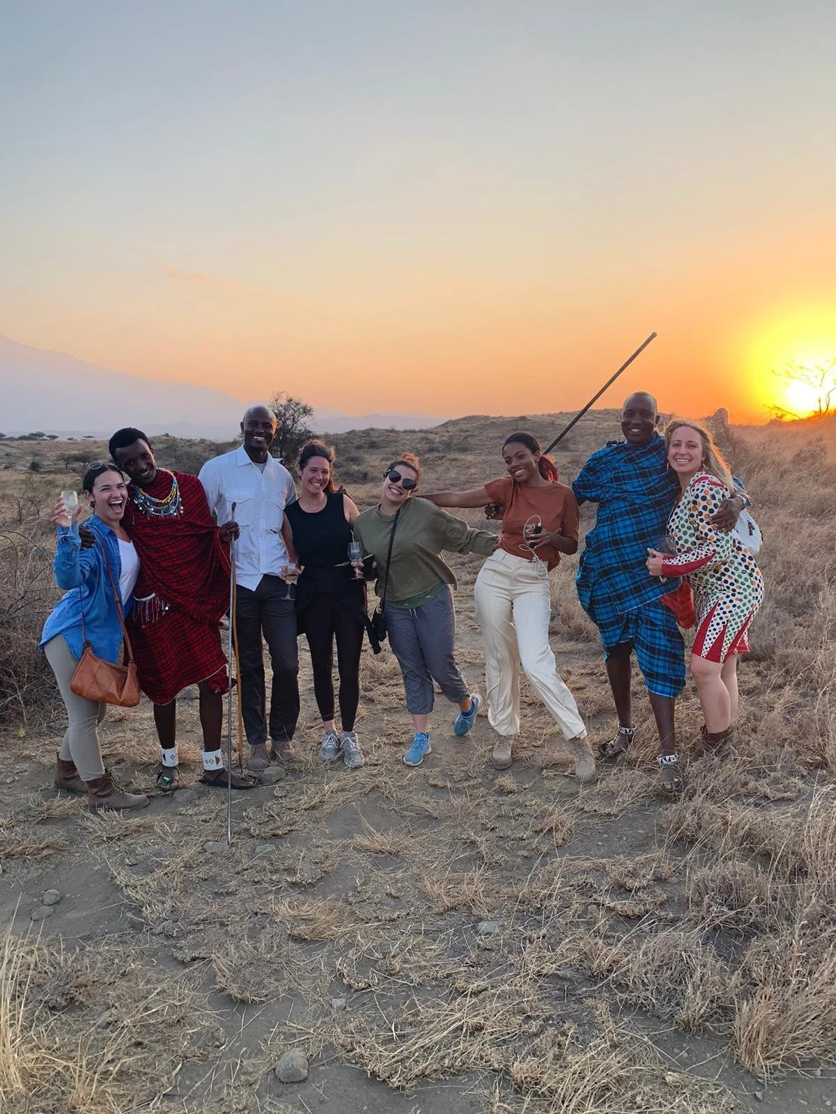 Group of eight people celebrating outdoors during sunset in a dry, grassy landscape, some holding glasses, and one person with a walking stick.