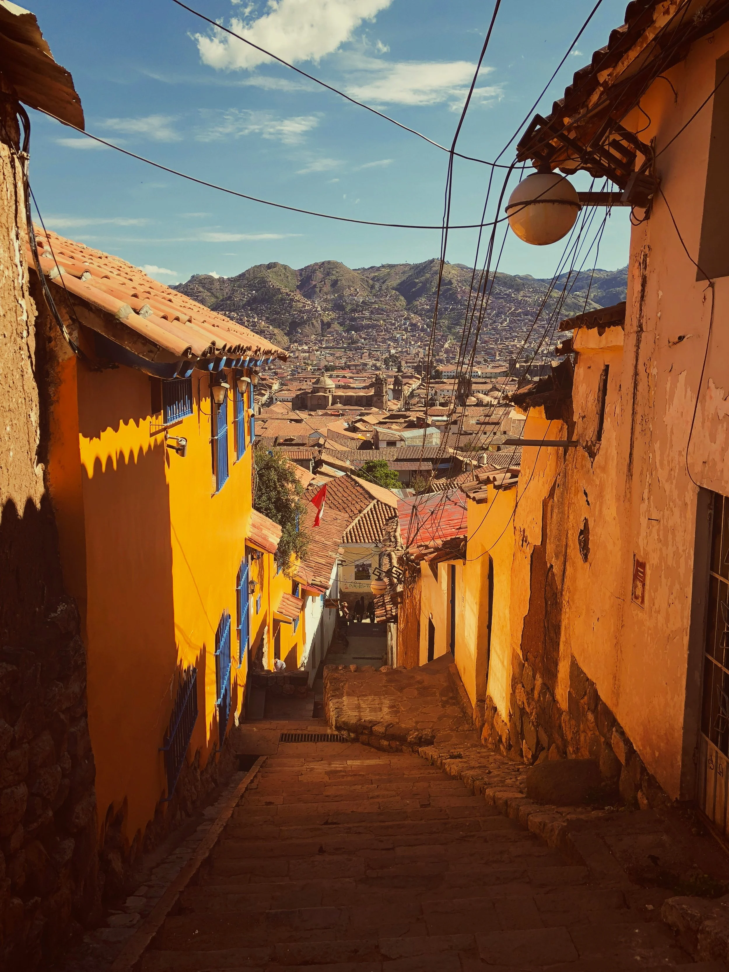 A narrow alleyway in a hilly town with colorful buildings on either side. The buildings are painted yellow and orange, with blue window frames. Overhead, electrical wires are strung across the alley. In the background, there are mountains and a sprawling city view under a partly cloudy sky.