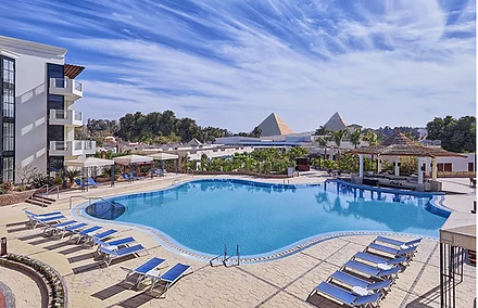 Swimming pool area in a resort with lounge chairs, a building on the left, and pyramidal structures in the background under a partly cloudy sky.