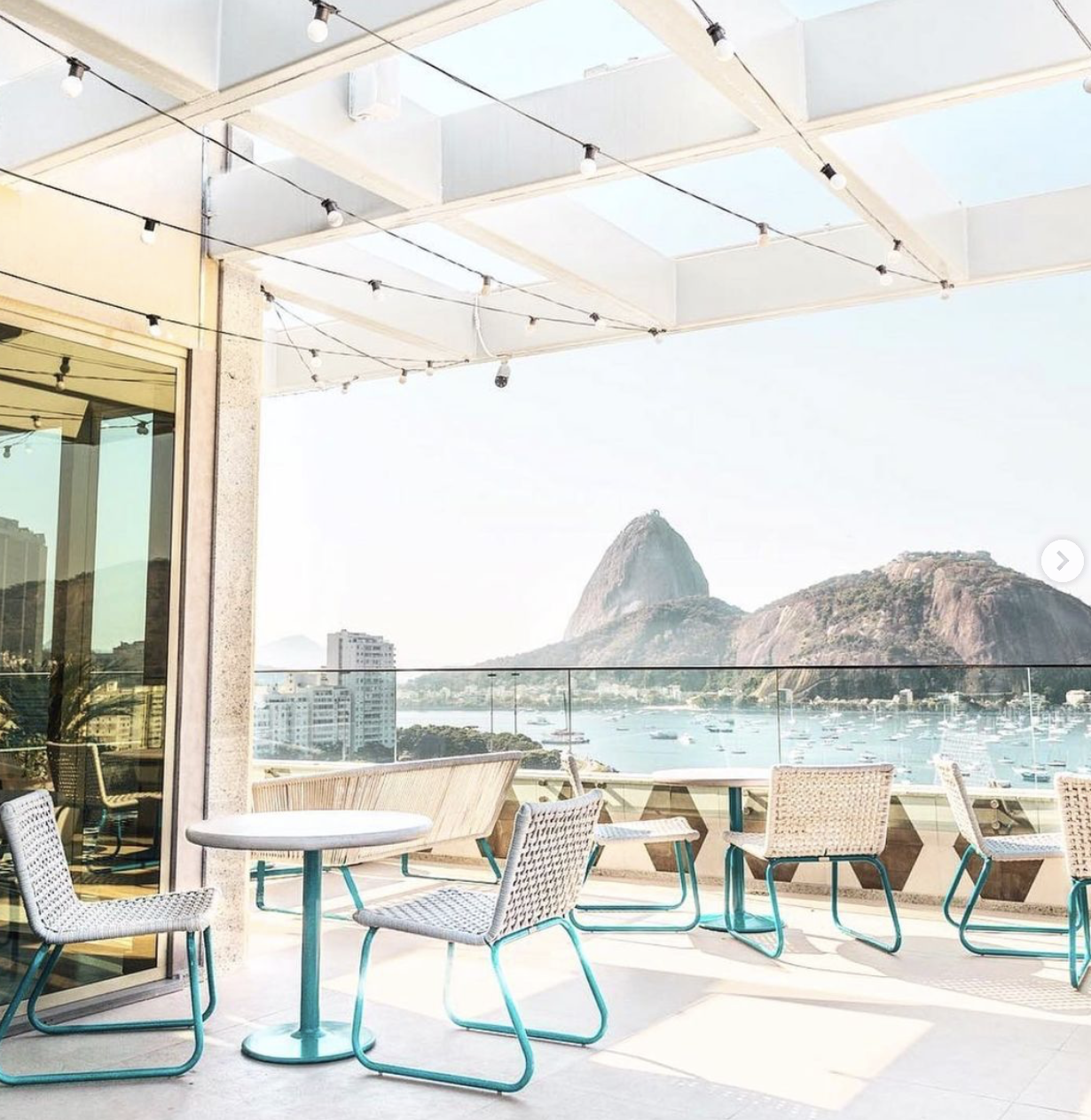 Outdoor terrace with tables and chairs overlooking a bay with boats and Sugarloaf Mountain in Rio de Janeiro, Brazil, under a partial glass roof with string lights.