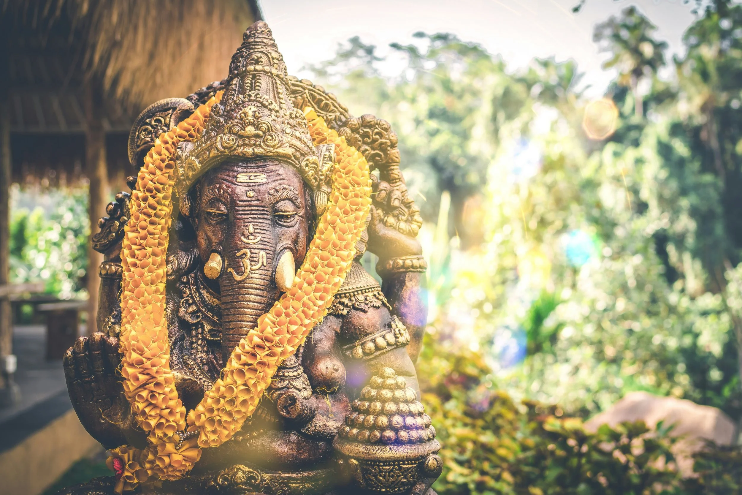A brass statue of the Hindu god Ganesha adorned with a yellow flower garland, set outdoors with blurred greenery in the background.