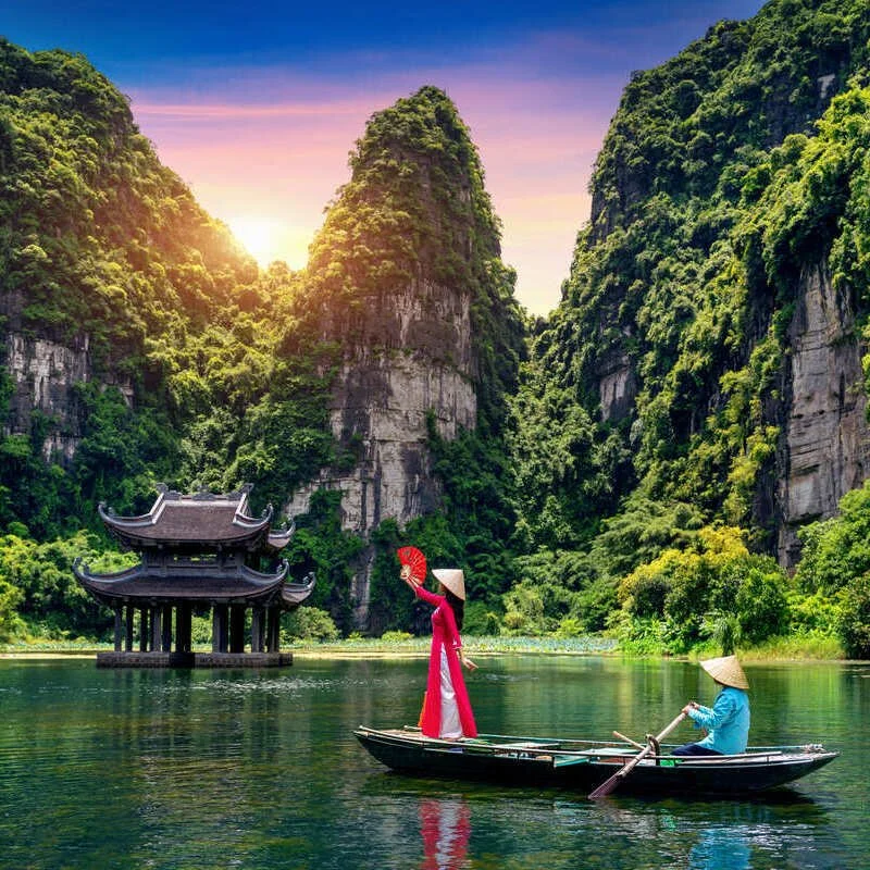 People in traditional Vietnamese clothing on a boat in a river surrounded by lush green cliffs, with a pagoda-style pavilion and a setting sun in the background.