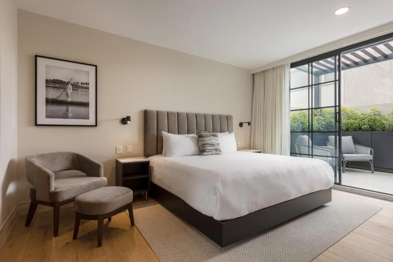 Modern hotel room with a large bed, beige walls, a black and white framed photograph, a gray chair with an ottoman, nightstands, and a sliding glass door leading to a balcony with outdoor seating and greenery.