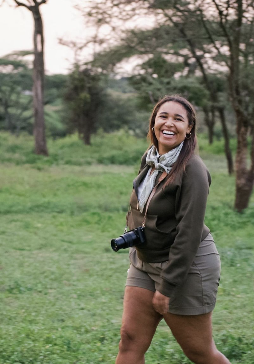A smiling woman with long brown hair, earrings, and a bandana around her neck, standing outdoors in a grassy area with trees, holding a camera.