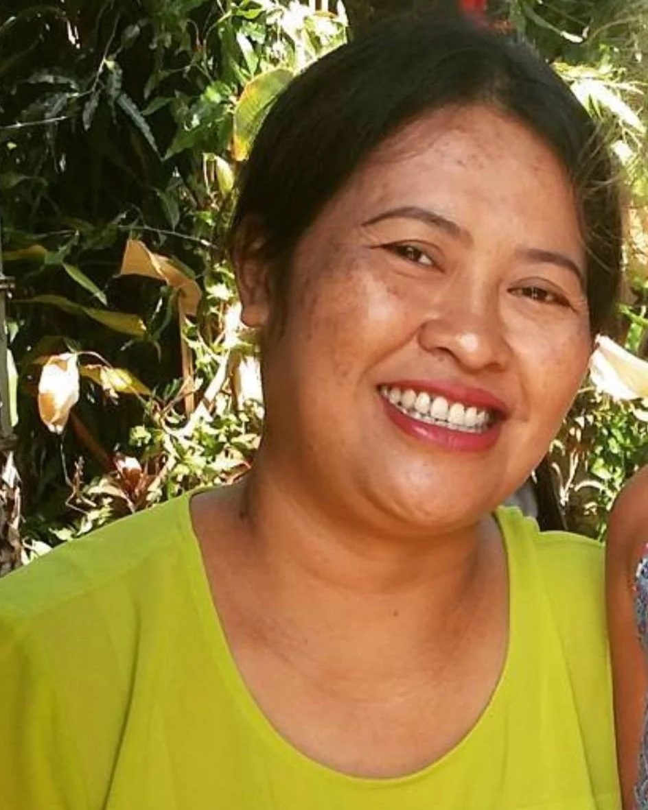 A woman with short black hair, wearing a bright green top, smiling outdoors with green foliage in the background.