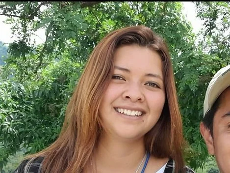 A young woman with long light brown hair smiling outdoors with green trees in the background.