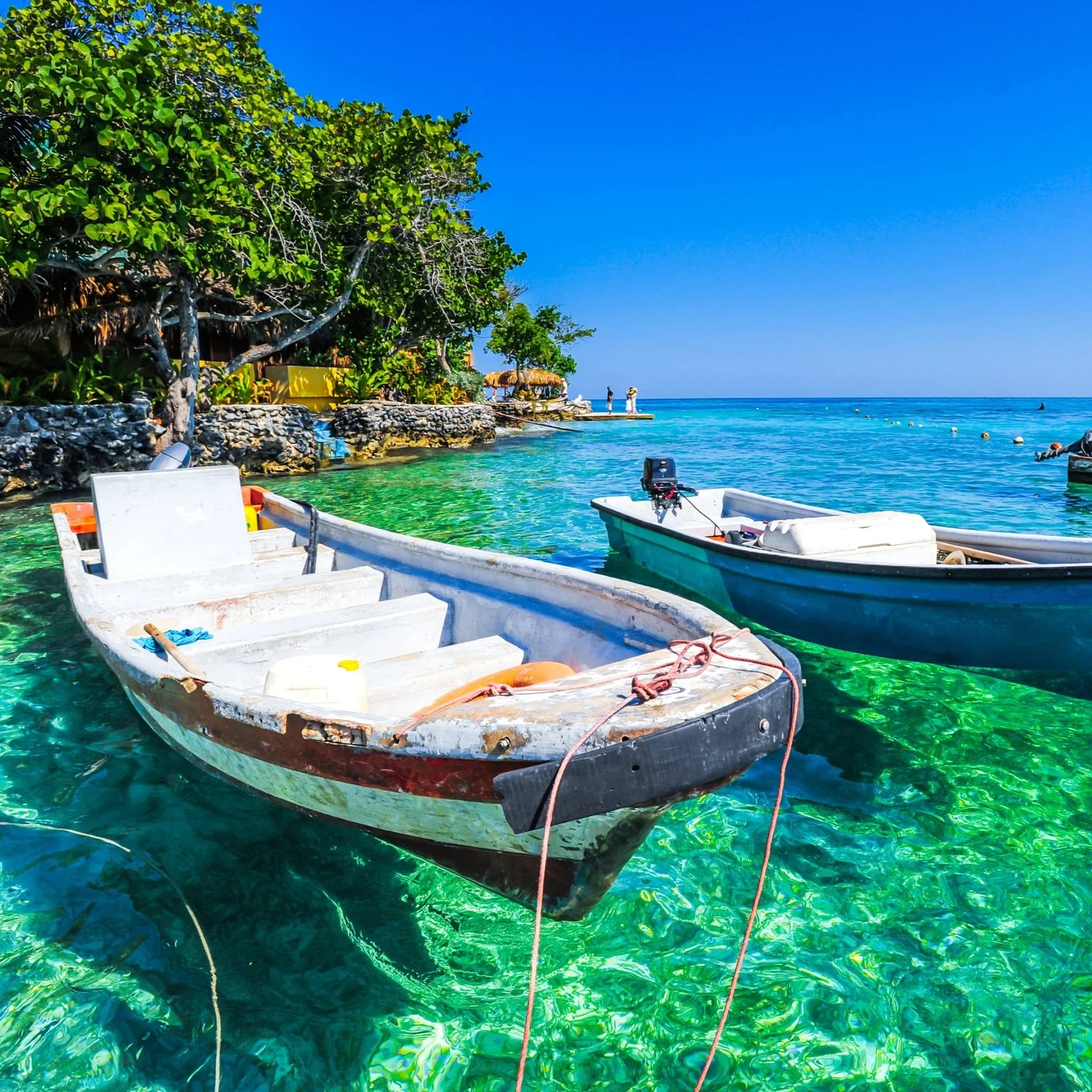 Two small boats floating on clear turquoise water near a rocky shoreline with trees and people in the background on a sunny day.