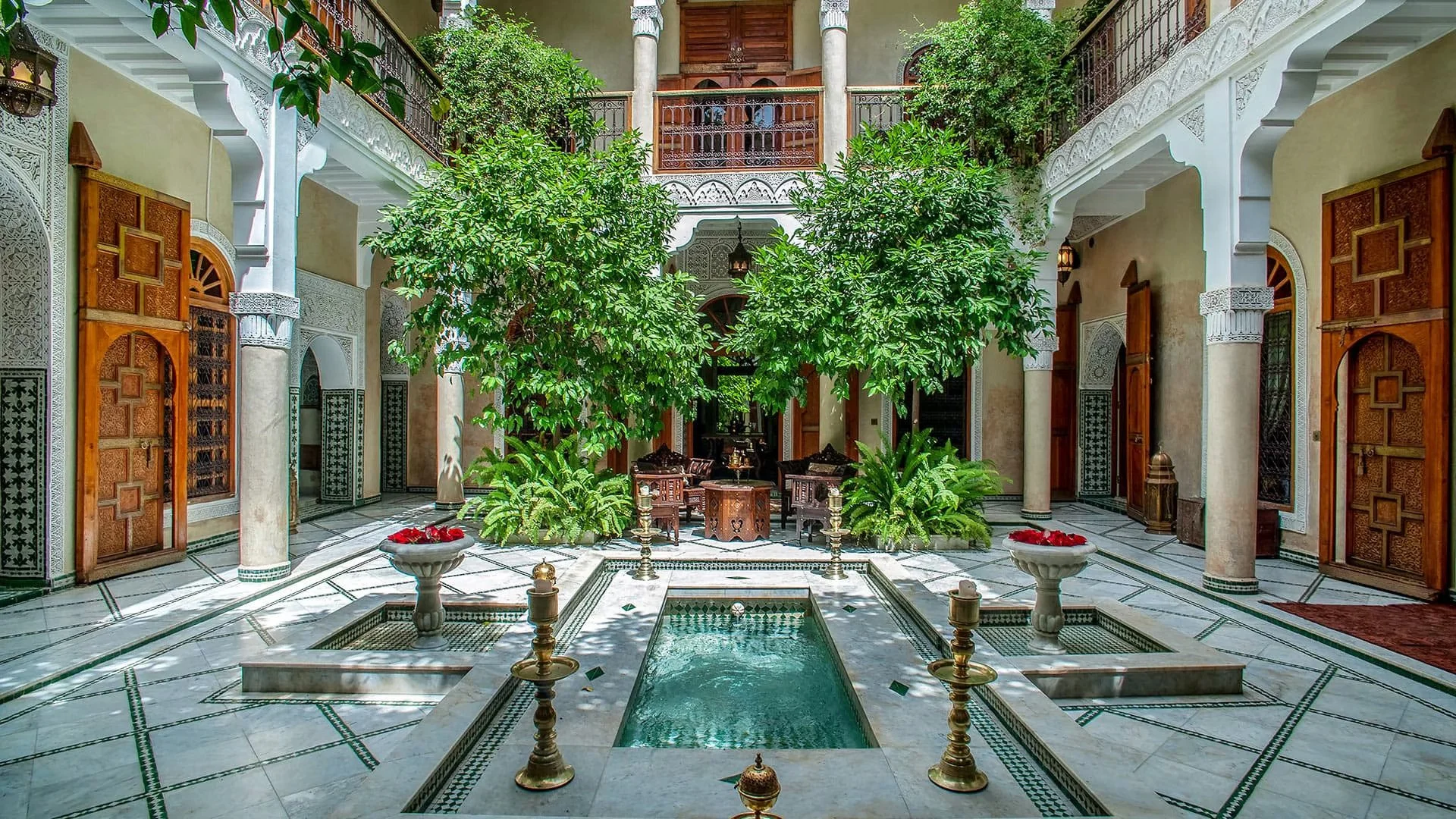 Interior view of a traditional Moroccan courtyard with a small central pool, lush green trees, ornate wooden doors, marble flooring, decorative tiles, and intricate white arches.