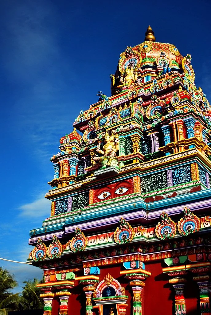Colorful Hindu temple with intricate carvings, statues, and large eyes painted on the front, set against a blue sky.