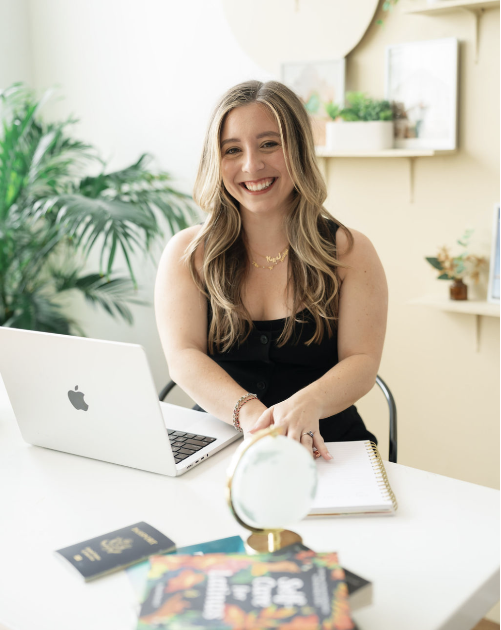 Young woman with long wavy hair sitting at a white desk, smiling, with a laptop, notebook, and travel books, in a bright room with houseplants and framed pictures.