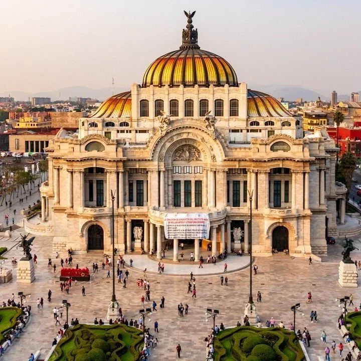 A large ornate building with a golden-tiled dome and decorative sculptures, surrounded by a plaza with people walking and greenery in Mexico City.