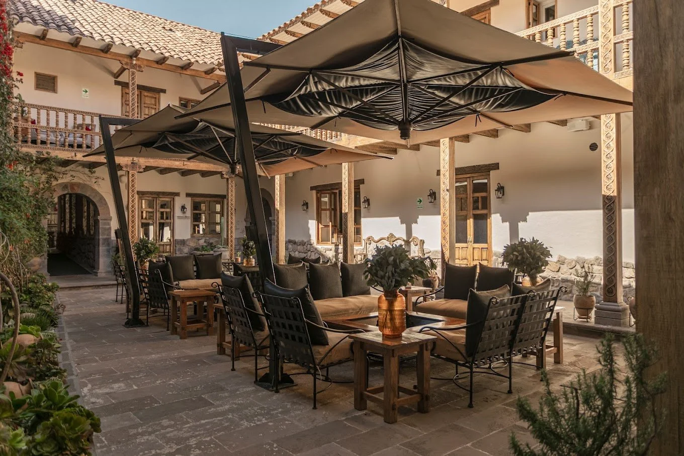 Outdoor patio area with wooden furniture, black chairs, and large beige umbrellas, surrounded by a building with wooden doors, windows, and decorative columns.