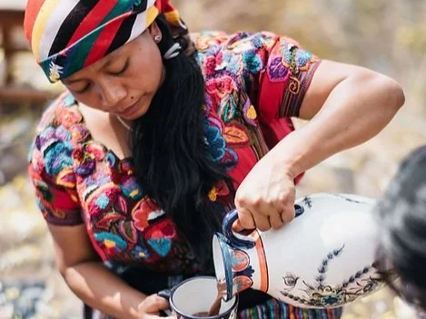 Woman wearing a colorful embroidered blouse and a striped headscarf pouring a beverage from a traditional ceramic pitcher.