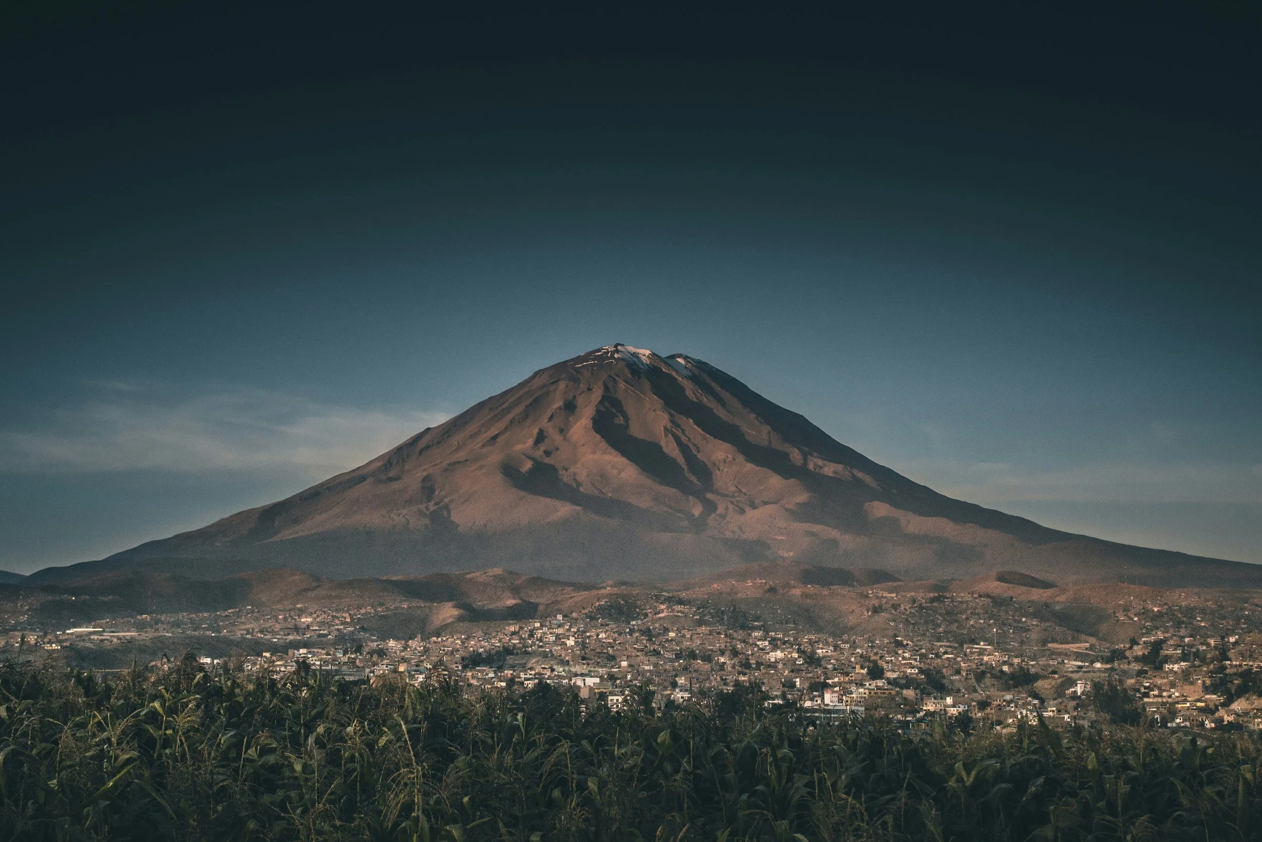A large mountain with a snow-capped peak overlooking a city and fields under a clear sky.
