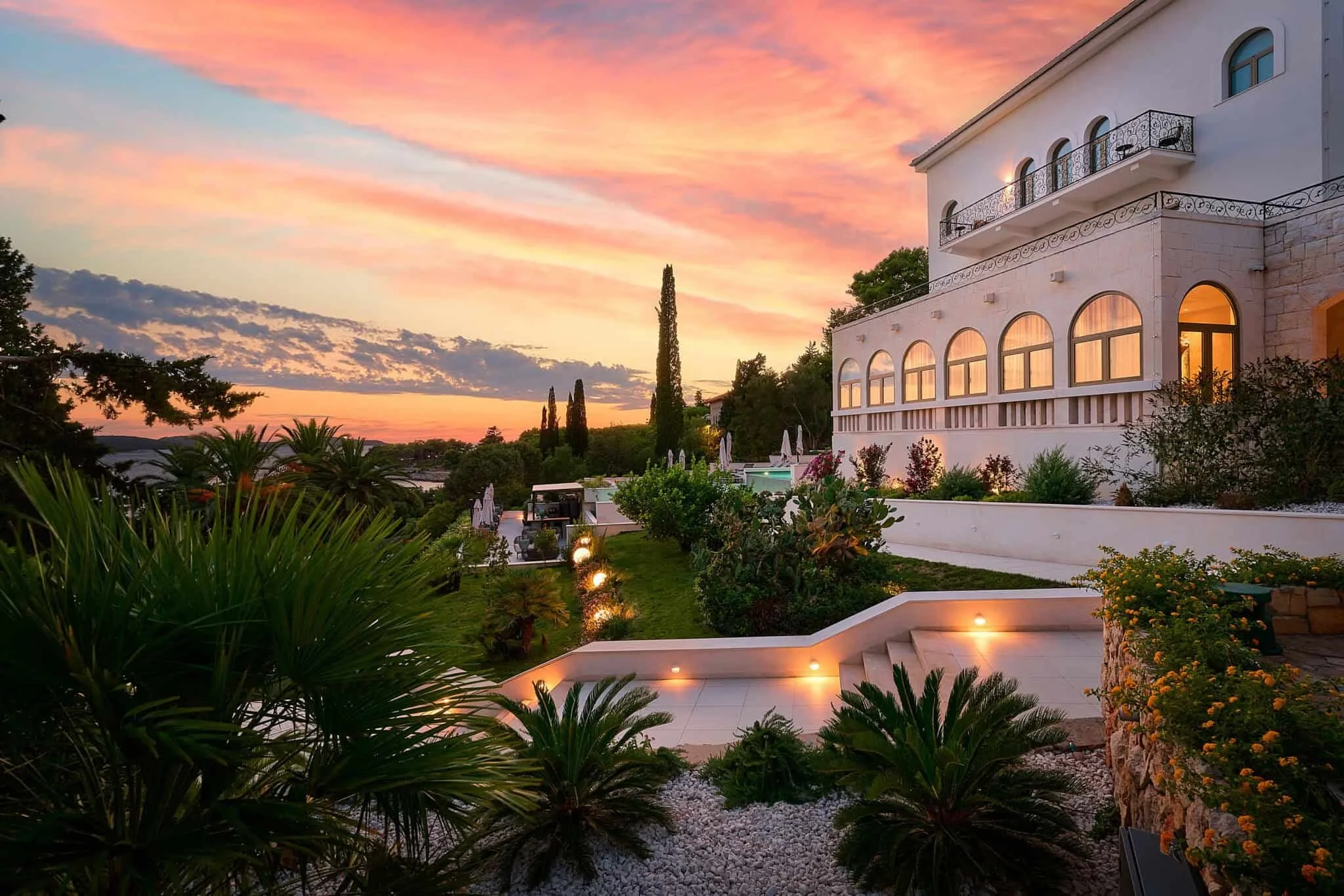 A large white Mediterranean-style villa with arched windows and a balcony, surrounded by lush greenery and garden lights at sunset.