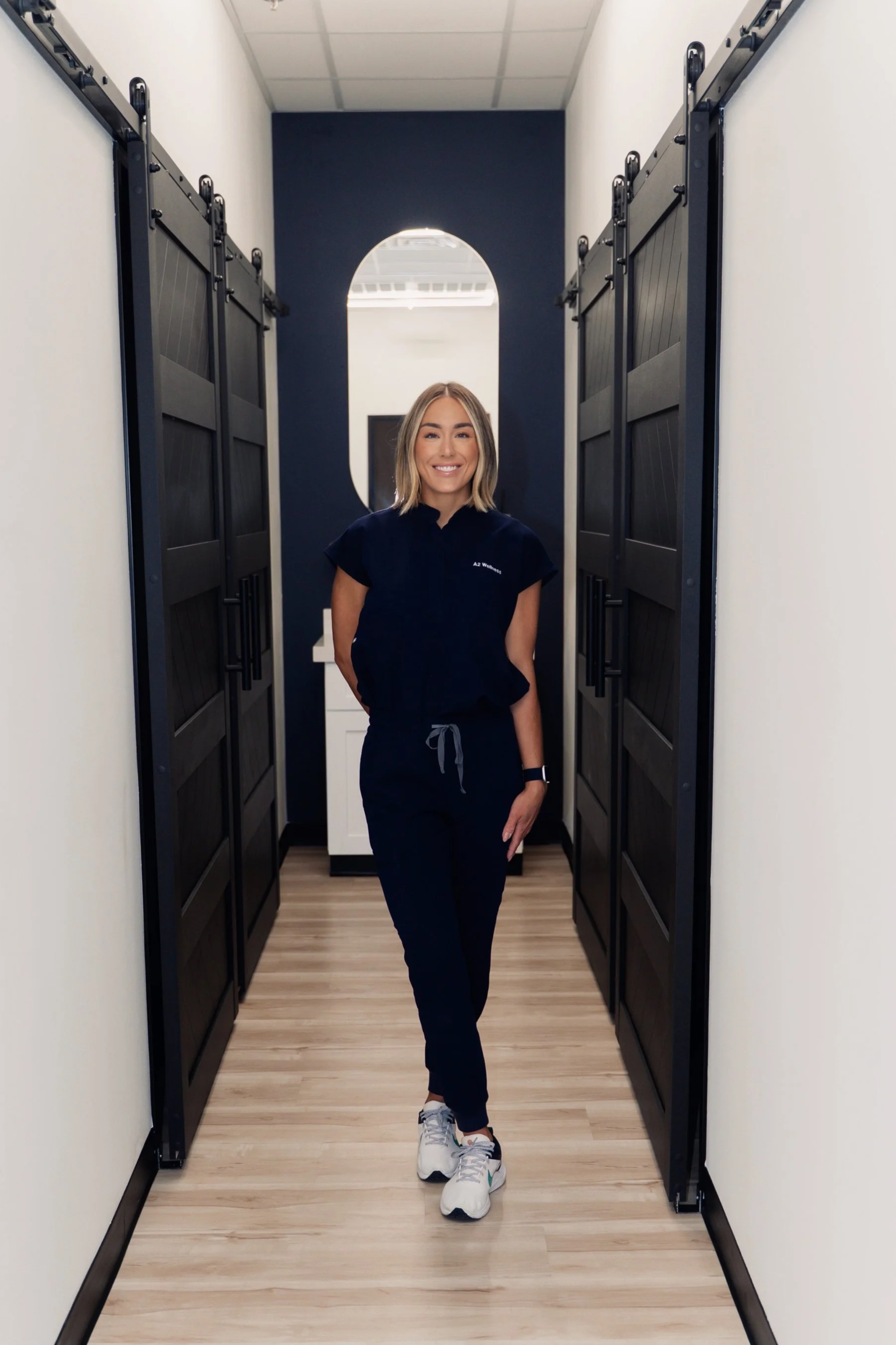 A smiling woman with shoulder-length blonde hair wearing a navy blue uniform, black pants, and white sneakers walking down a hallway with black sliding barn doors on both sides, white walls, and a wood floor.
