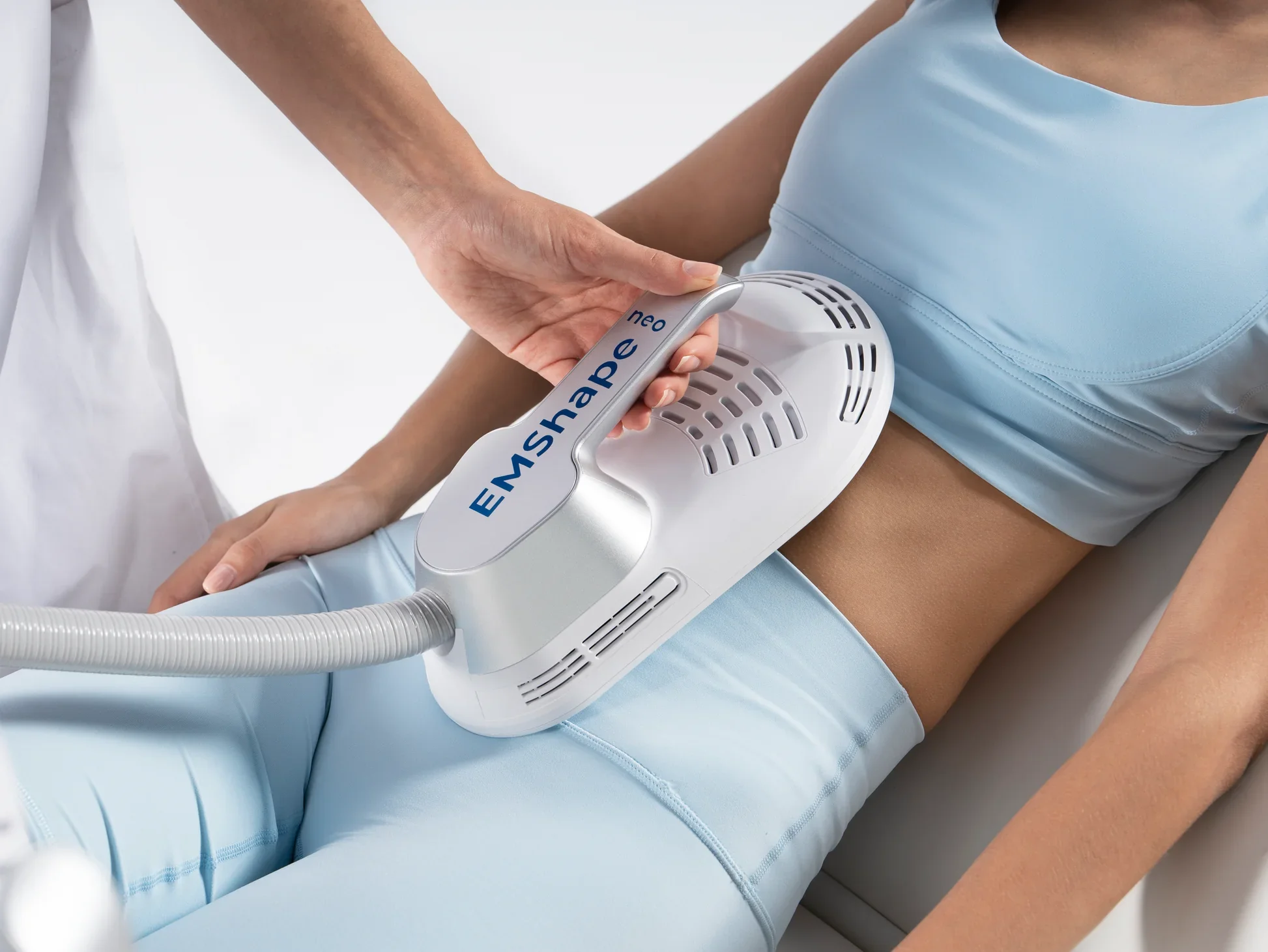 A medical procedure involving a woman in a blue top and matching pants receiving treatment with a device labeled EMShape Neo on her lower abdomen, while a healthcare professional operates the device.