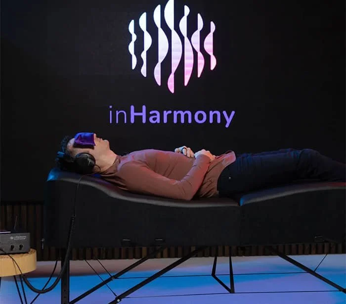Person lying on a lounge mattress with a VR headset in a room with a black background and the 'inHarmony' logo projected on the wall.