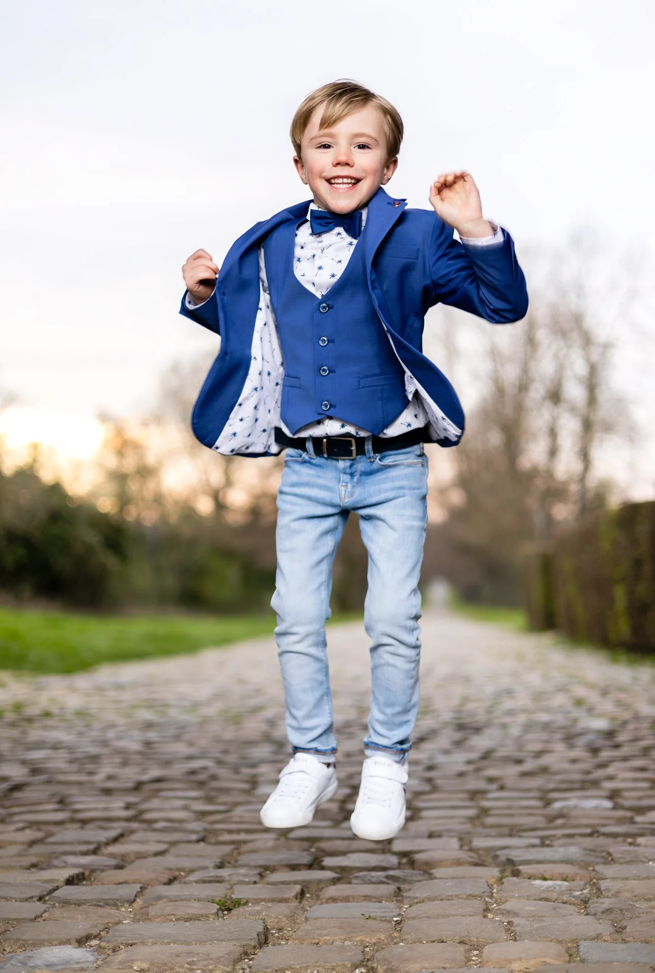 A young boy in a blue suit jacket, vest, and bow tie jumping on a cobblestone path outdoors with a happy expression.