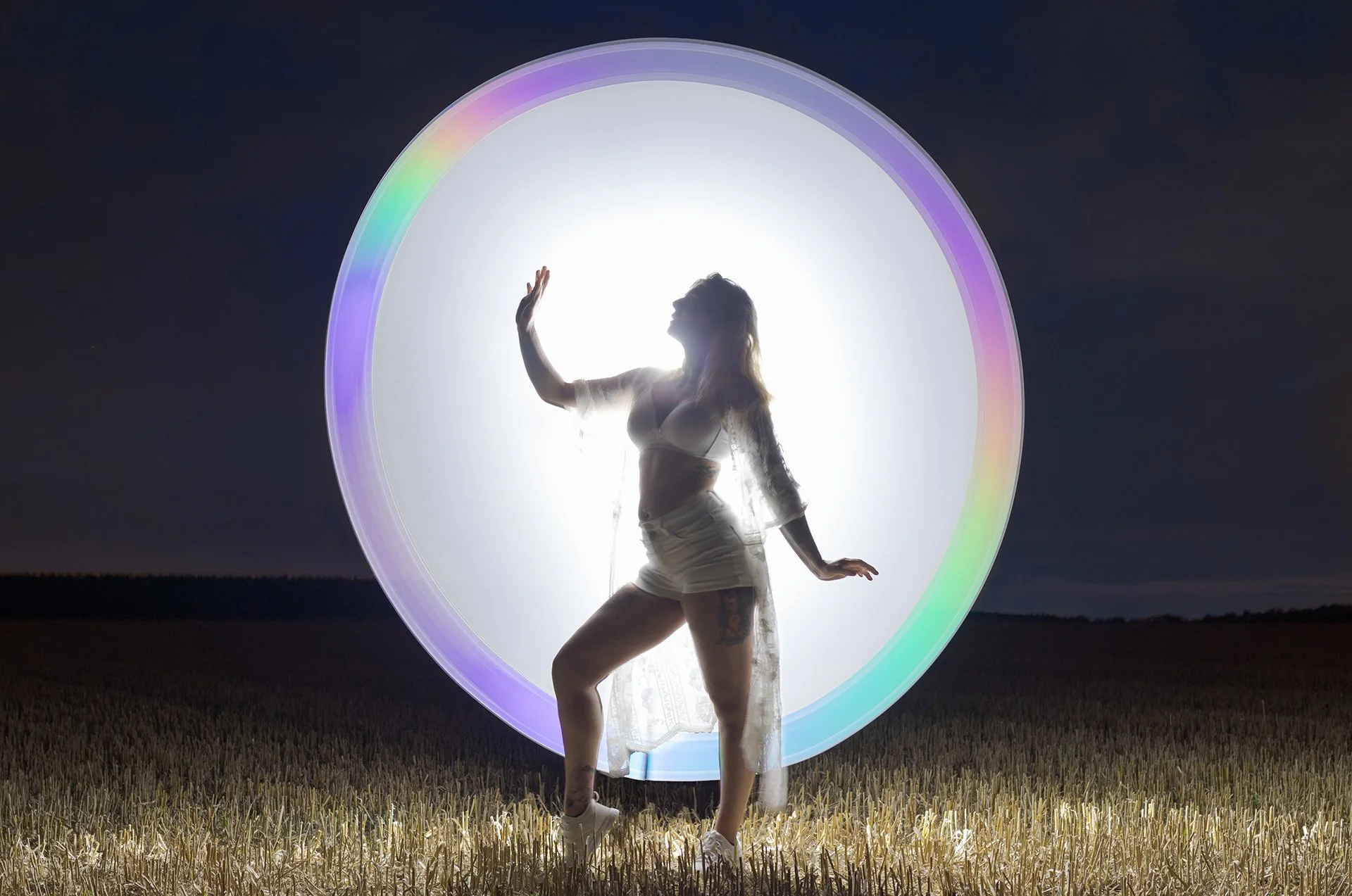 A woman dancing in a field at night, backlit by a large illuminated circular light with rainbow-colored edges.