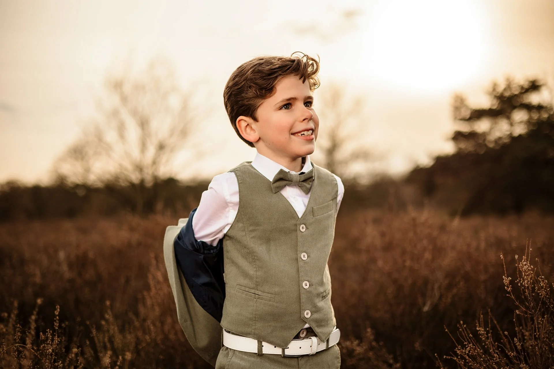 Communie photoshoot of a young boy dressed in a beige vest, white shirt, and matching beige pants, standing outdoors; smiling as he looks to the side, with a blurred background of trees and a warm sky.