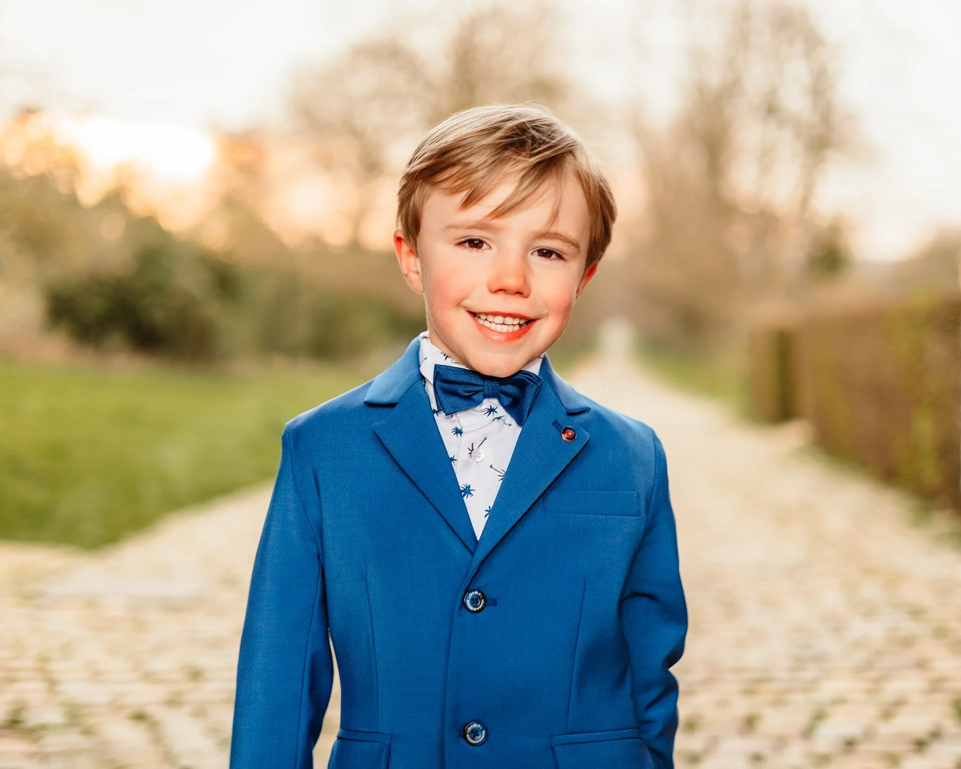 A young boy in a blue suit with a bow tie, smiling outdoors on a fall day with trees and a pathway in the background.