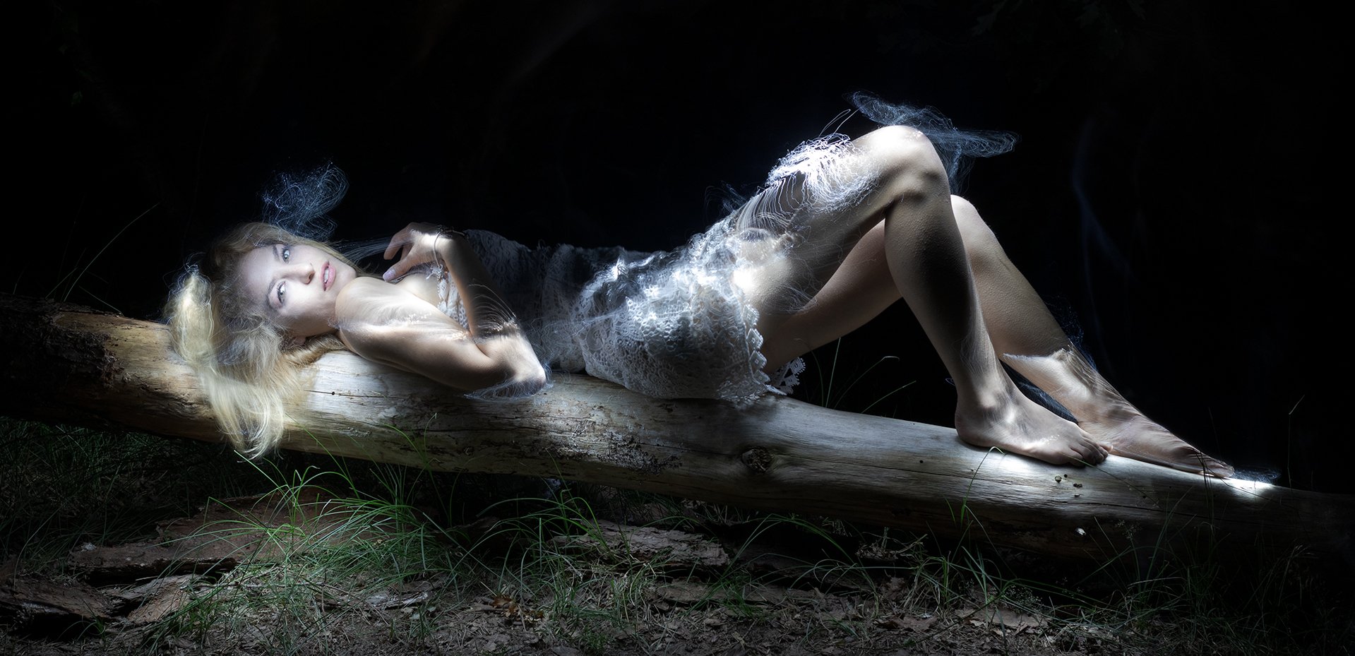A woman with blonde hair lying on a fallen log in a dark outdoor setting, illuminated by a dramatic light that highlights her face, hair, and body, creating an ethereal effect.