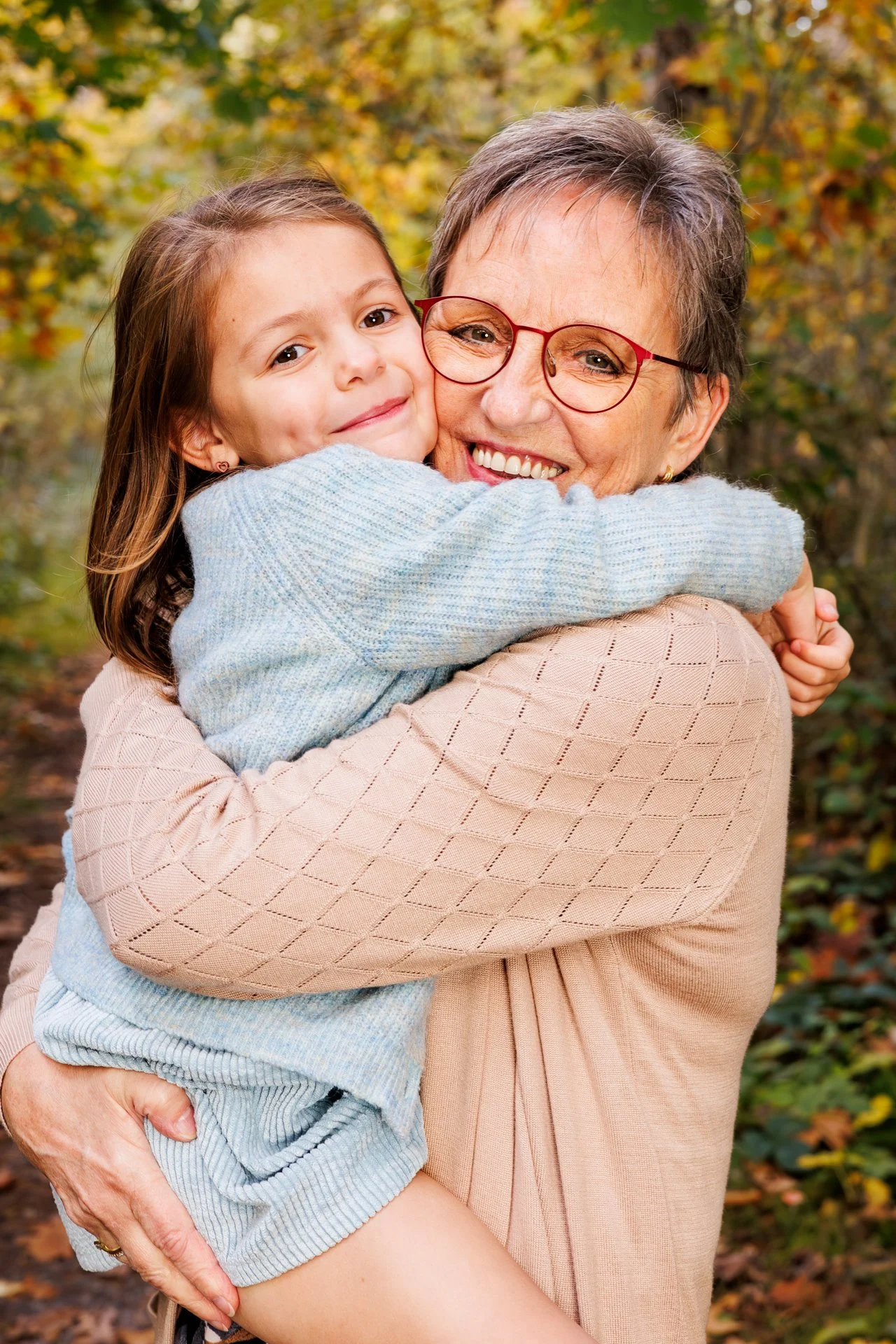 A young girl hugging an older woman, both smiling happily outdoors during autumn, with colorful fall trees in the background.