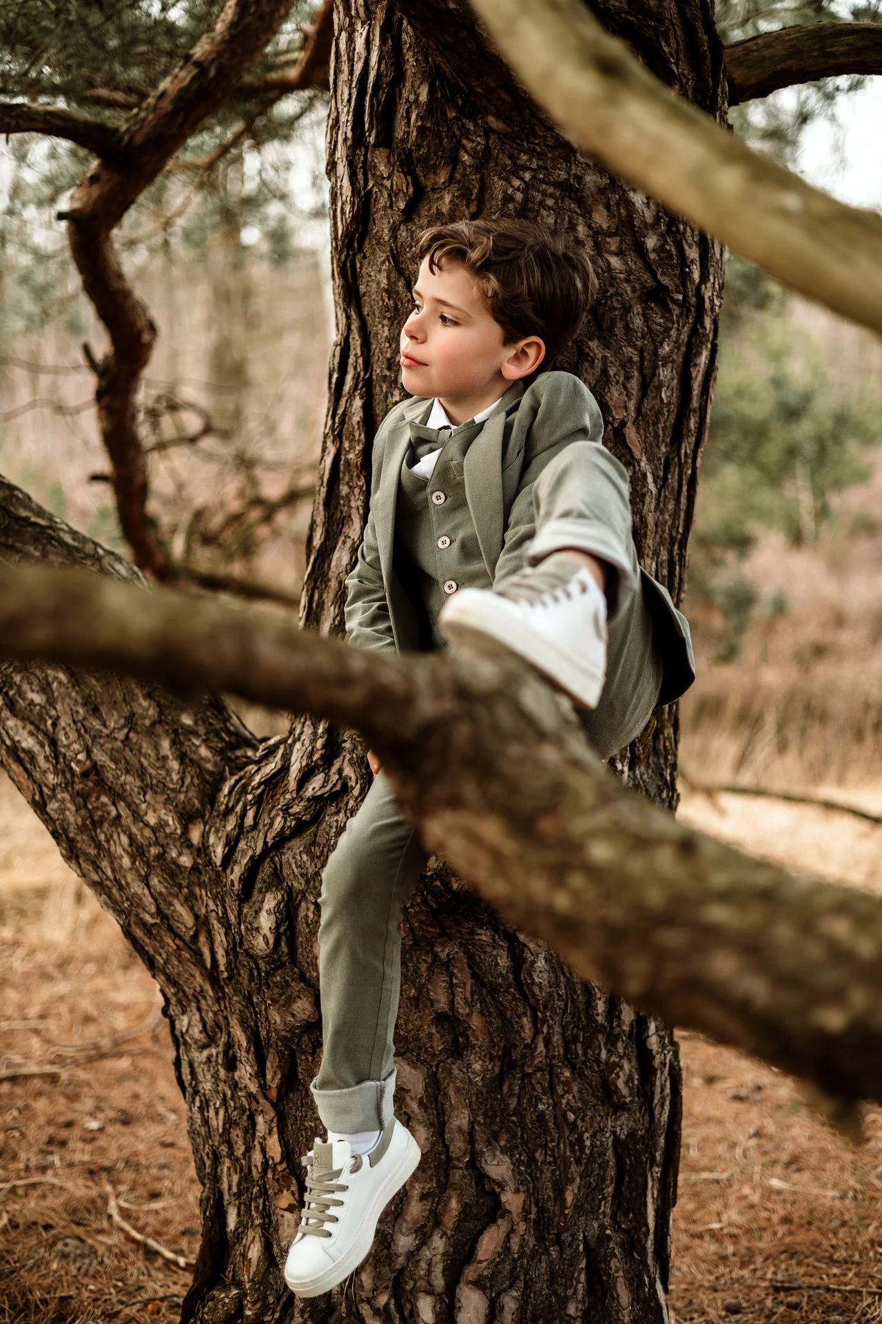 A young boy dressed in a green suit with sneakers, sitting on a tree branch in a wooded area.