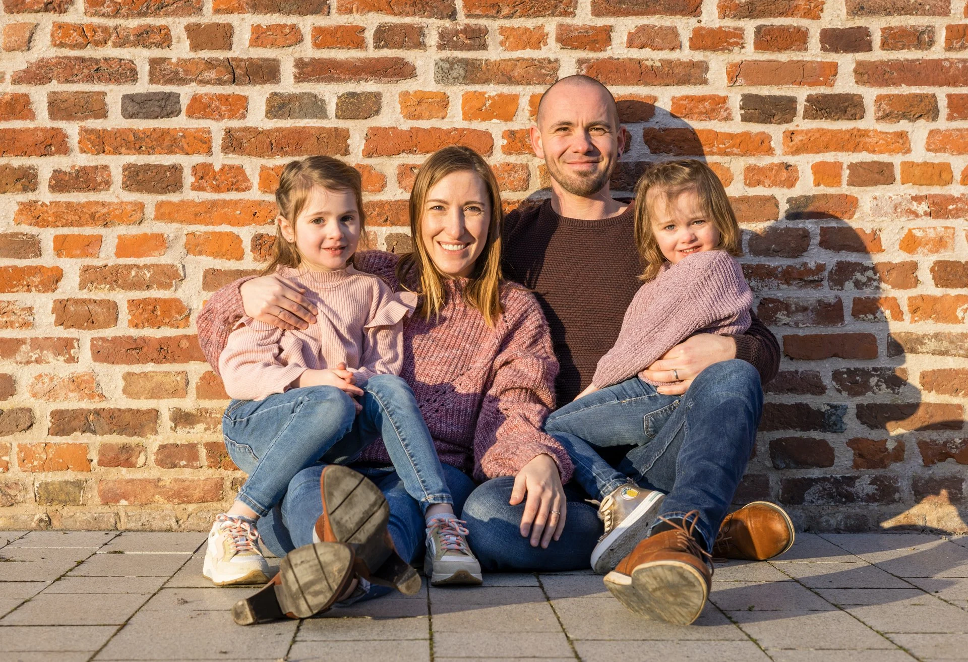 A family photoshoot of four sitting in front of a brick wall, smiling at the camera during sunset. The mother and father are seated with two young girls, all wearing casual clothing and sneakers.