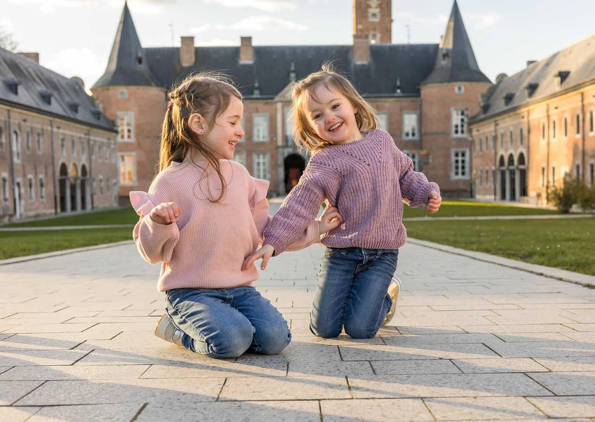 Family photoshoot of two young girls with light brown hair, wearing pink and purple sweaters, kneeling on a paved courtyard, smiling and playing together in front of a historic castle with brick walls and towers, during daylight.