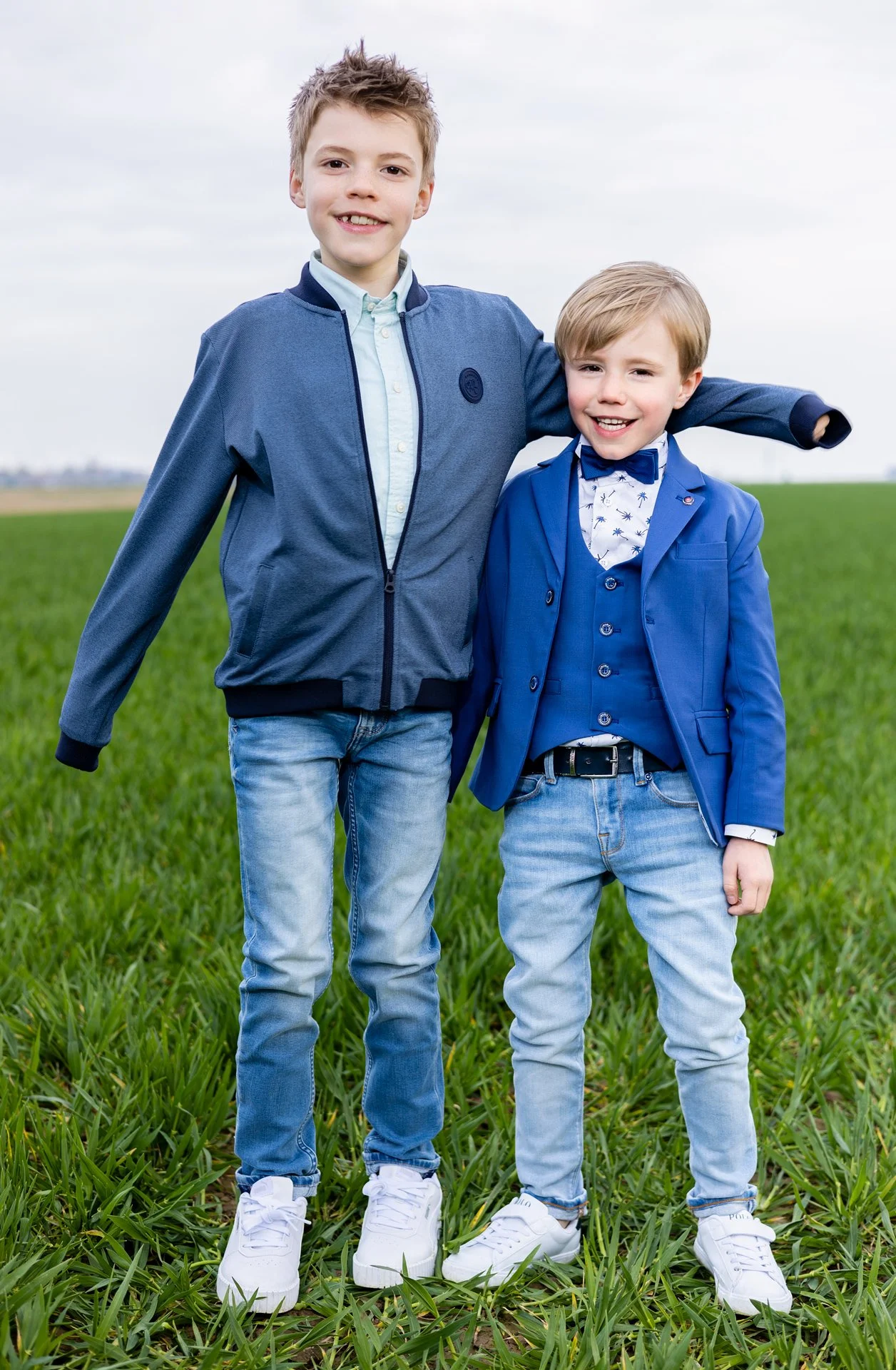 Two boys standing on a grassy field, smiling at the camera, with one boy's arm around the other's shoulder. They are dressed casually, with one in a blue jacket and jeans, and the other in a blue blazer and dress shirt. The sky is overcast in the bac