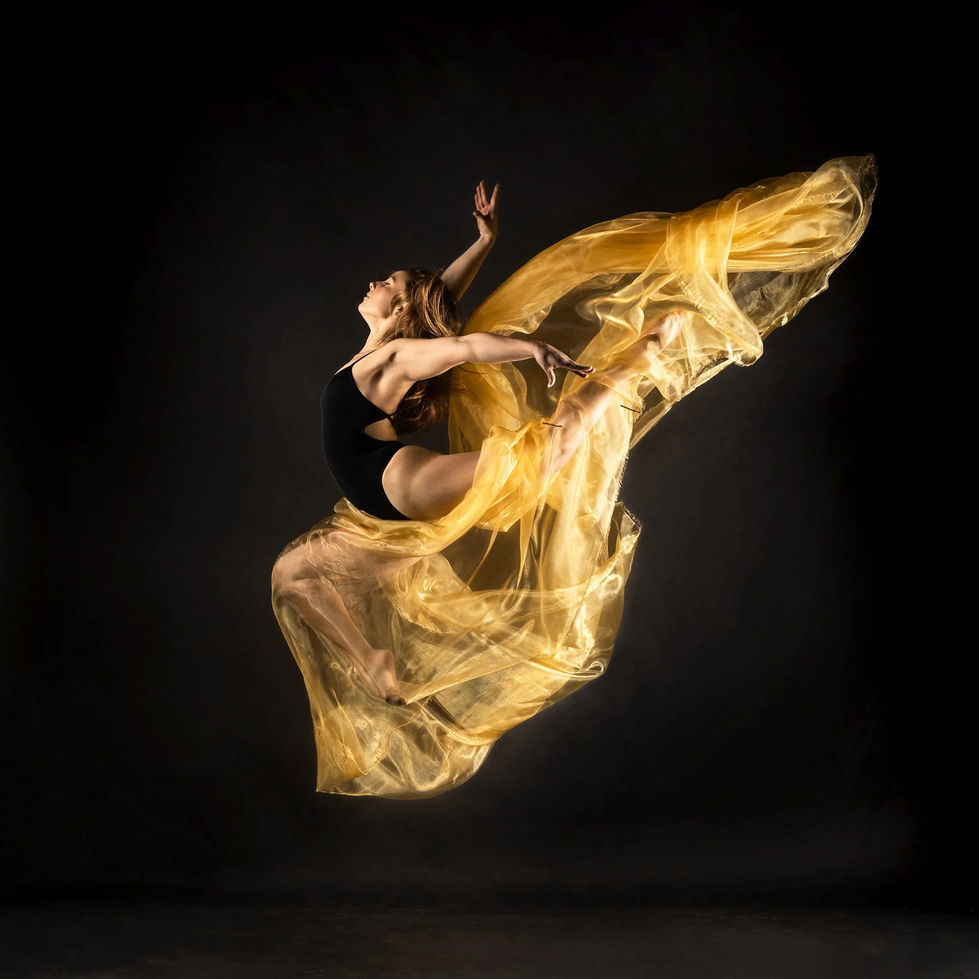 A female athlete in black dancewear performing an energetic leap with flowing yellow fabric against a black background.