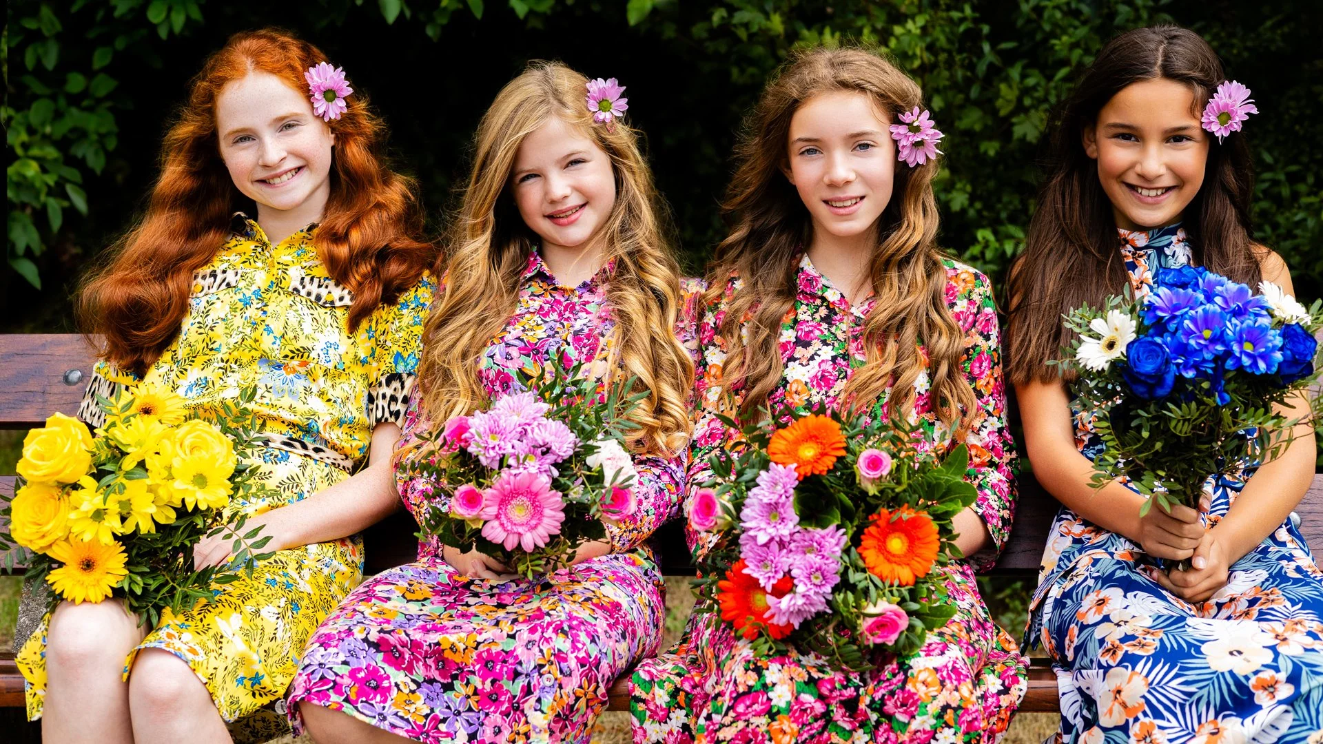 Four young girls sitting on a park bench, each holding colorful bouquets of flowers, wearing patterned dresses with flowers in their hair.