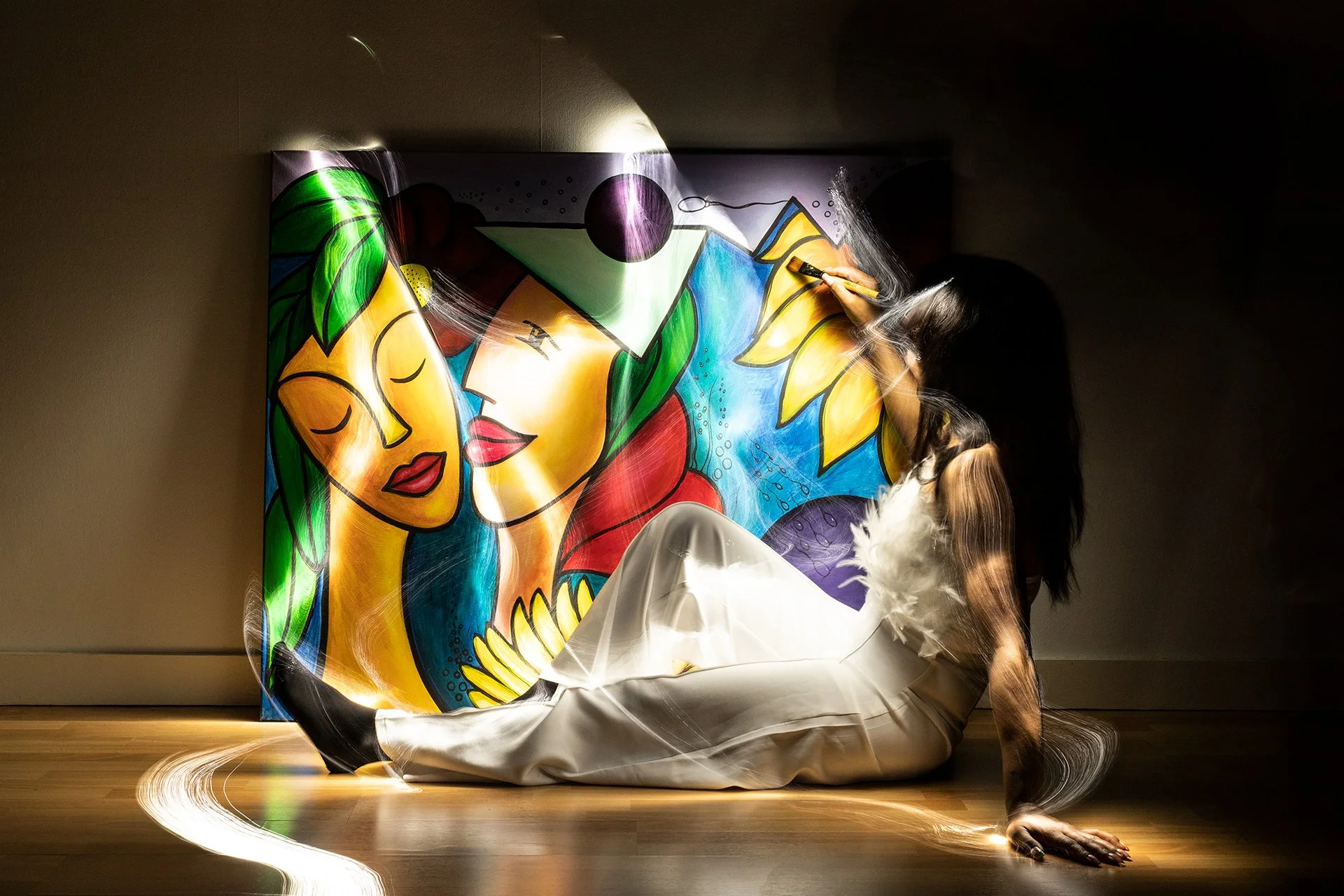 A woman sitting on the floor in front of a colorful abstract mural, with lighting and motion effects highlighting her hair and clothing with light painting.