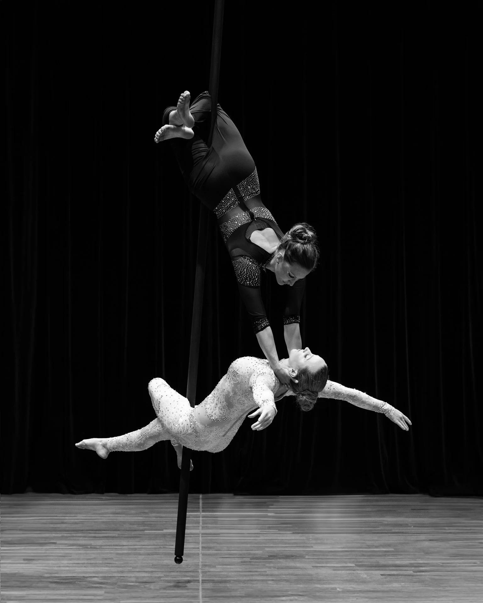 Two women performing an aerial acrobatic routine on a silk, with one woman upside down and the other supporting her, on a stage with dark curtains.