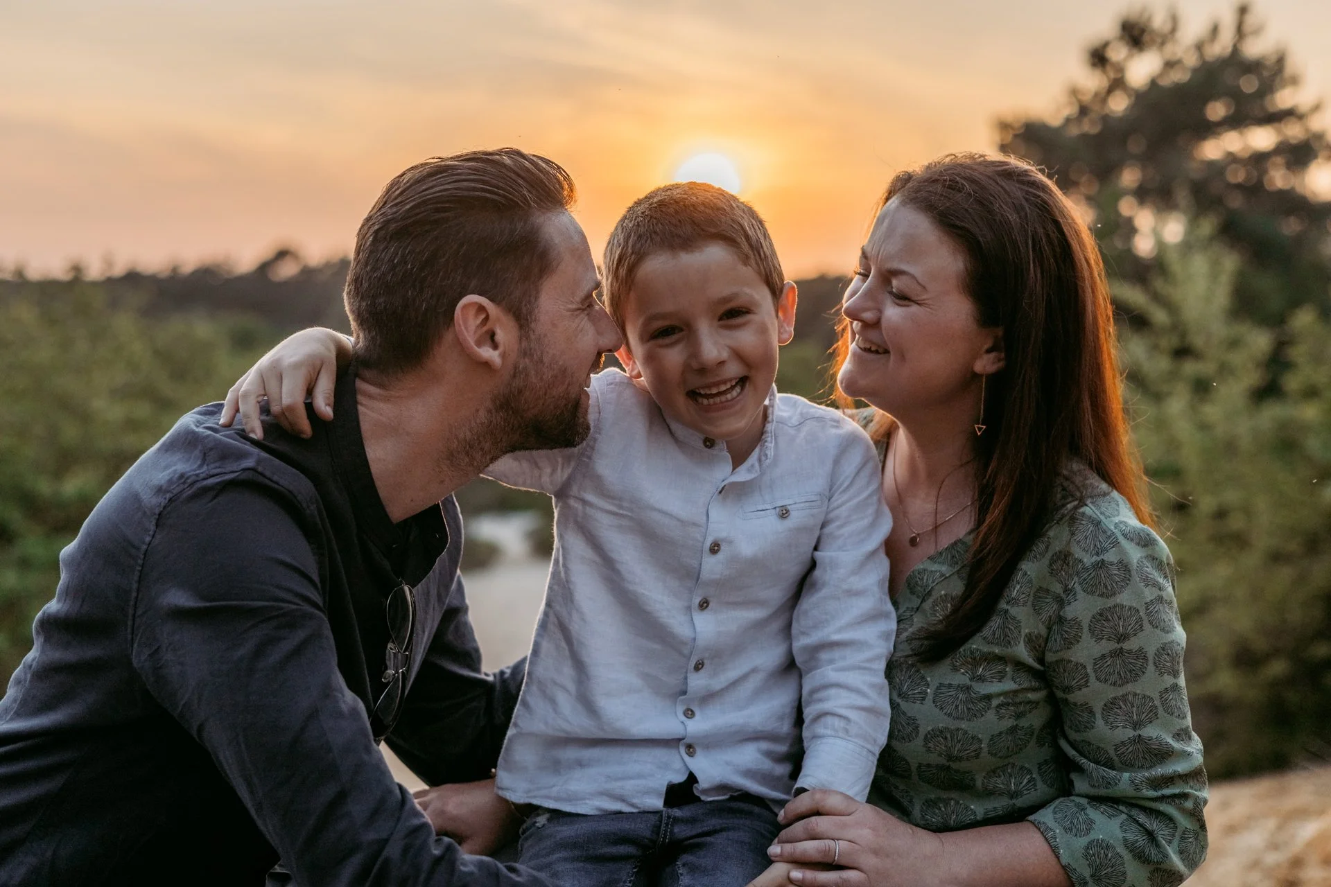 A young boy and his parents smiling and laughing outdoors at sunset, with the boy sitting on his mother's lap and his father leaning in close.
