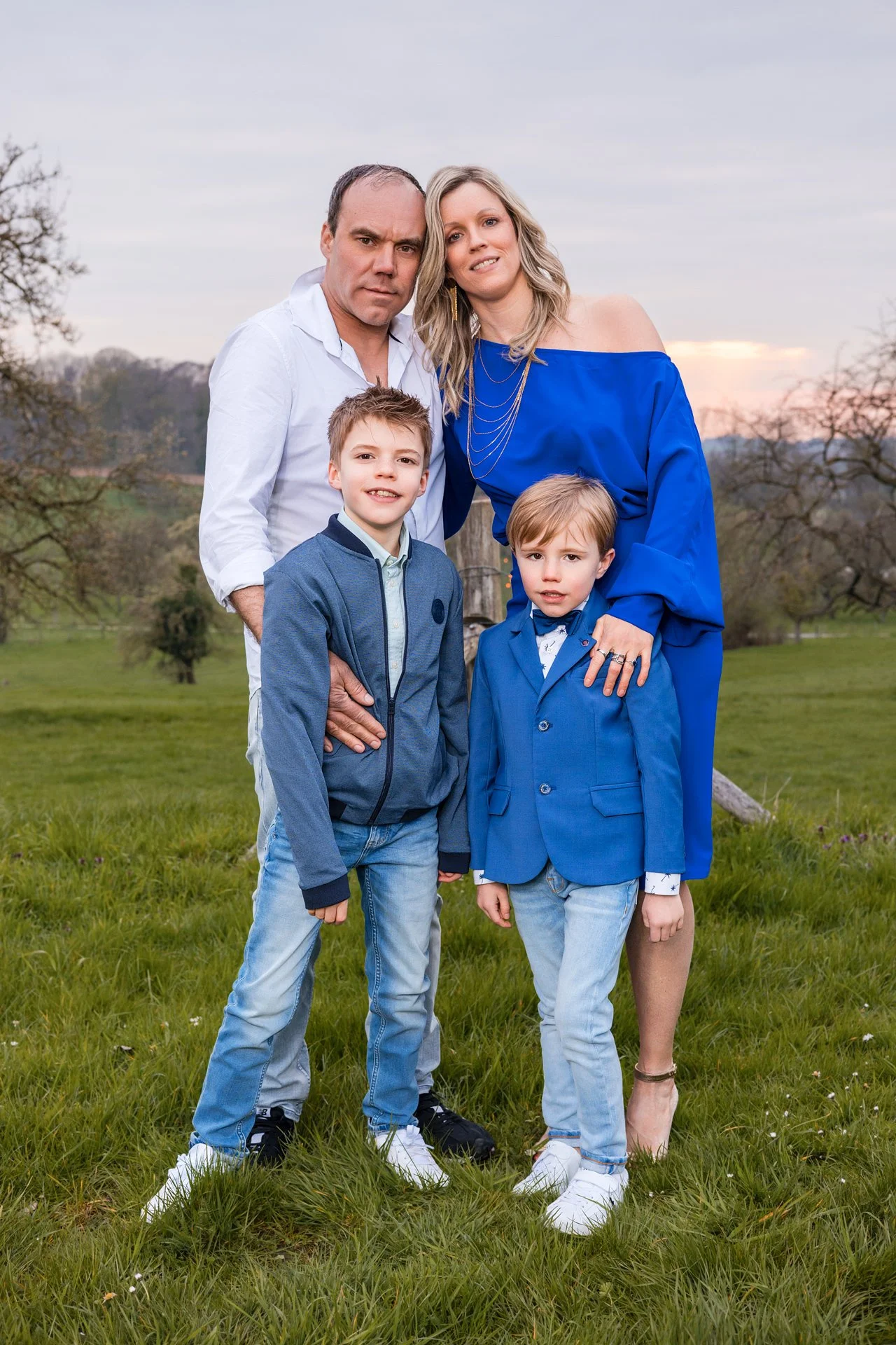 A family of five outdoor in a grassy field during sunset, with trees in the background. The father is in a white shirt, the mother in a blue dress, and two young boys in blue jackets and jeans. They are standing close together, smiling and posing for