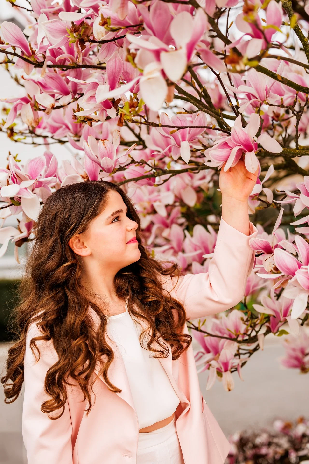 A woman with long, curly brown hair wearing a light pink blazer and white top standing next to a magnolia tree in bloom with pink and white flowers.