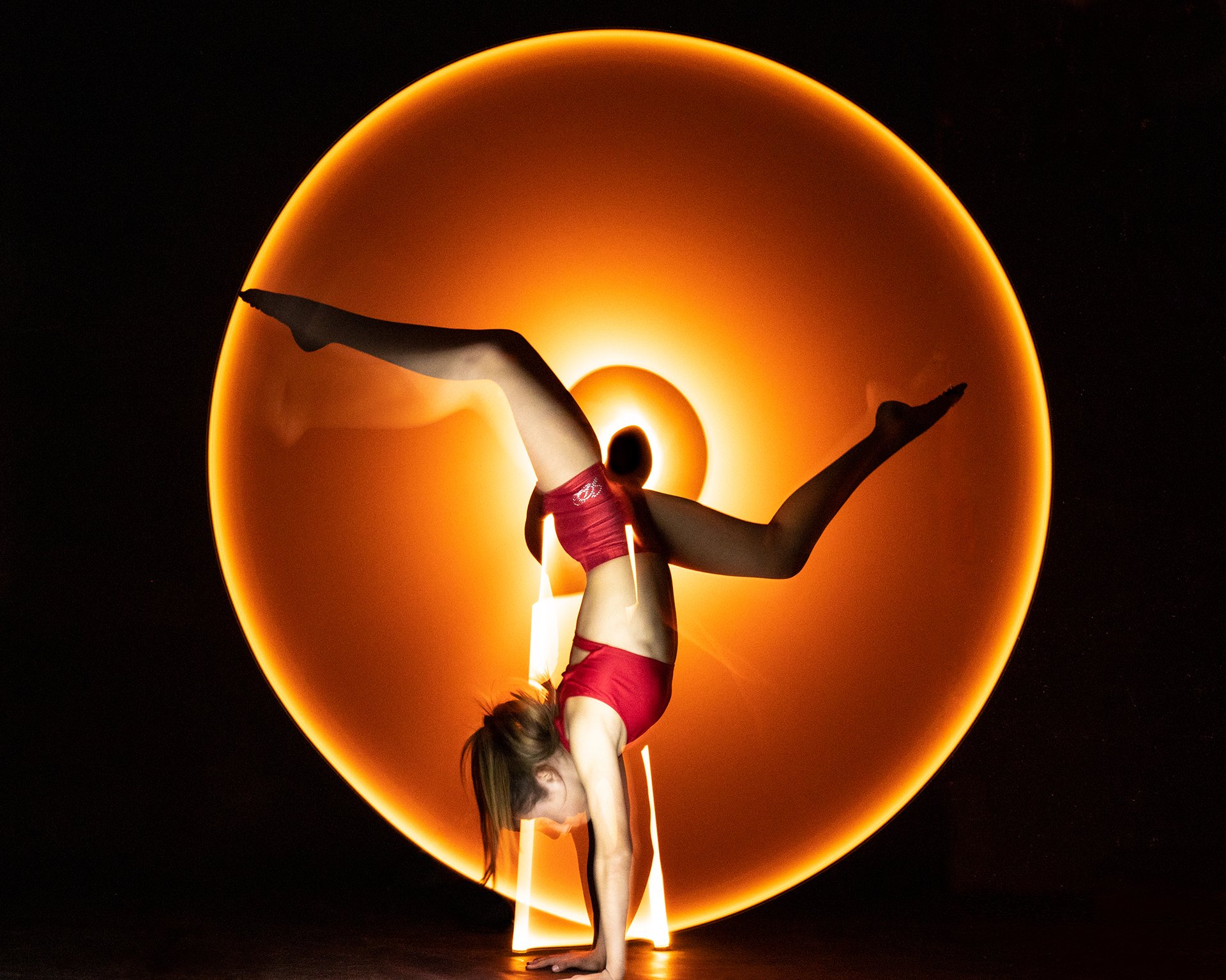A woman practicing a handstand in front of a large, orange, glowing circular light. Light painting.